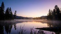 Serene mountain lake at dawn with misty water surface, evergreen forest silhouettes and purple sunrise sky creating peaceful wilderness landscape.