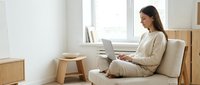 Young woman working on laptop computer while sitting in comfortable beige armchair near window in modern minimalist living room with wooden furniture.