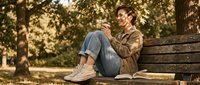 Young Caucasian man in casual autumn outfit sitting on wooden park bench reading book during golden hour sunlight filtering through trees.