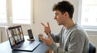 Young Caucasian man in gray sweater having animated video conference call on laptop, gesturing while speaking to colleagues on screen in bright office.
