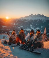 Four snowboarders sitting together on snowy mountain slope during golden hour sunset with alpine peaks in background for winter sports and friendship concepts.