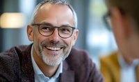 Mature Caucasian businessman with gray hair and glasses smiling during meeting, wearing brown sweater. Professional corporate portrait showing confidence and success.