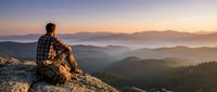 Young Caucasian male hiker sitting on rocky mountain peak, enjoying breathtaking sunset over misty valley, concept of freedom and adventure.