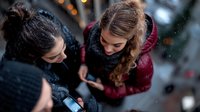 Young diverse women in winter clothing looking at mobile phones during snowfall, sharing social media content outdoors in evening city lights atmosphere.