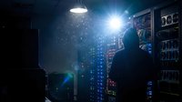 Young male technician in dark uniform inspecting server equipment in dimly lit data center with glowing blue lights and network hardware visible on shelves.