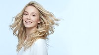 Young blonde Caucasian woman with flowing hair in motion wearing white top smiling joyfully against light blue background for beauty and lifestyle content.