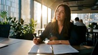 Professional Asian businesswoman writing in notebook at modern office desk with natural sunlight streaming through windows during productive workday.