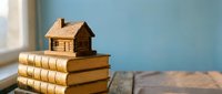 Wooden miniature house model sitting on stack of old books against blue wall background representing real estate education and property investment concepts.