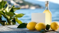Fresh lemons and homemade limoncello bottle with blank label on wooden surface against blurred Mediterranean coastline background with green leaves.