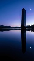 Tall dark obelisk monument reflected in still lake water at twilight with purple sky, moon, and mountains in background for architectural and landscape projects.