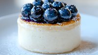 Fresh blueberries topped on creamy white cheesecake dusted with powdered sugar, close up view on light background showing texture and layers of dessert.