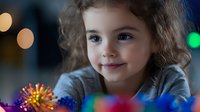 Young caucasian girl with curly hair smiling while lying down, surrounded by colorful Christmas lights creating magical bokeh effect in dark background. Holiday portrait.