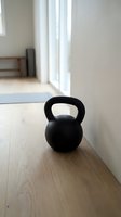 Black kettlebell weight on wooden floor in bright, minimalist gym studio with natural light from windows, suitable for fitness, training, and wellness content.