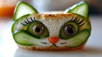 Creative sandwich art featuring cucumber slices forming cat ears and eyes, with olive pupils and tomato nose on bread, showcasing playful food presentation.