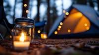 Glowing mason jar lantern and illuminated tent at twilight campsite with string lights creating magical outdoor atmosphere in forest setting.