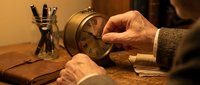 Businessman hands adjusting vintage brass alarm clock on wooden desk with pen holder in warm office lighting for time management and productivity concepts.