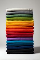 Stack of neatly folded colorful textile fabrics arranged in rainbow spectrum from white to black, showcasing vibrant hues for home decor or fashion design.