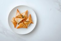 Golden crispy phyllo triangles filled with savory ingredients served on white plate against marble background, overhead view of Mediterranean appetizers.