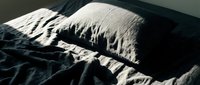 Unmade bed with white pillow and dark rumpled sheets, morning sunlight casting dramatic shadows across wrinkled fabric and bedding.