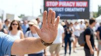 Activist raising hand at public demonstration against privacy violations and government overreach with crowd and protest signs in background.