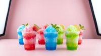 Colorful layered slushie drinks with ice and fruit garnishes in clear cups against pink background, suitable for beverage menus, summer refreshment marketing, and food photography.