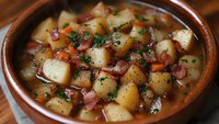 Hearty potato soup with tender chunks of potato, diced carrots, and smoky ham in rich broth, garnished with fresh herbs. Served in rustic ceramic bowl.