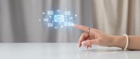 Hand touching digital email and communication icons floating above white desk surface with blurred office background, representing online messaging and digital correspondence.