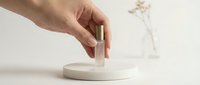 Hand placing a small glass perfume bottle with gold cap on a white marble podium, minimal aesthetic, soft light, beauty product presentation.