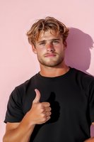 Young Caucasian male with wavy blonde hair wearing black t-shirt shows thumbs up against pink wall. Strong shadow creates dramatic contrast for modern lifestyle portrait.