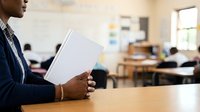 Male teacher of African descent holding blank papers in classroom with students at desks, suitable for education and training materials.