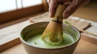Matcha tea preparation with traditional bamboo whisk in ceramic bowl, whisking green powder into hot water for ceremonial beverage making.
