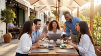 Young diverse professionals gathered around outdoor dining table with laptop, reviewing work documents during lunch break in sunny patio setting with umbrellas and greenery.
