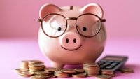 Pink ceramic piggy bank wearing glasses with stacked coins and calculator on pink background, representing smart financial planning and savings concept.
