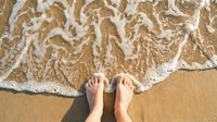 Bare feet standing on golden sandy beach with foamy ocean waves washing ashore creating relaxing vacation atmosphere for summer travel concepts.