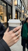 Female hand holding disposable coffee cup on urban street with storefronts and pedestrians blurred in background, suitable for beverage, lifestyle, and city living concepts.