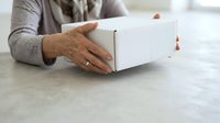 Senior woman with aged hands opening white cardboard box on marble surface, wearing gray sweater. Suitable for e-commerce, shipping, delivery, and unboxing concepts.