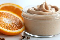 Creamy beige whipped body butter or skincare mousse in glass jar next to fresh orange slice and clove spices on white background.