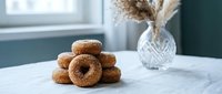 Fresh glazed donuts stacked on marble counter in modern kitchen with natural window light and decorative pampas grass vase creating cozy breakfast scene.