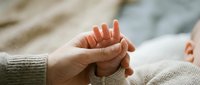 Adult hands gently holding tiny newborn baby fingers in tender moment of connection and love between parent and child in soft natural light.