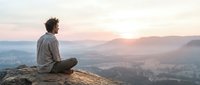 Man with curly hair sitting on rocky cliff edge overlooking misty valley landscape at sunrise, contemplating nature and peaceful morning scenery.