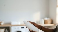 Hand holding white cosmetic bottle with brown liquid product in bright minimalist bathroom setting with natural light and wooden accessories.