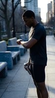 Young African American man checking smartwatch during urban morning jog in city street with skyscrapers and golden sunlight for fitness tracking.