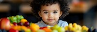 Young mixed-race toddler with curly hair sitting at kitchen table surrounded by fresh colorful fruits and vegetables for healthy eating concept.