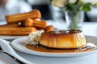 Golden caramel flan dessert with glossy sauce on white plate, traditional custard pudding served in elegant kitchen setting with flowers in background.