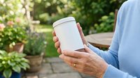 Mature woman holding blank white container in garden setting with green plants and flowers in background, suitable for product mockups and lifestyle marketing.