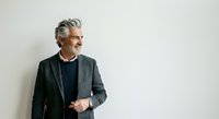 Mature man with gray hair and facial hair, wearing dark blazer over navy shirt, standing against white background, looking to side with confident expression, suitable for corporate and professional business use.