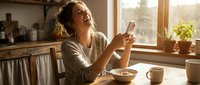 Woman with curly blonde hair in casual beige cardigan sitting at wooden table by sunny window, holding smartphone, enjoying breakfast with coffee and bowl. Warm home interior setting.
