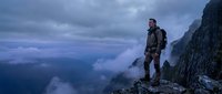 Male hiker in tactical gear standing on mountain cliff edge overlooking misty valley during dramatic cloudy weather conditions for outdoor adventure concepts.