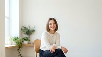 Young woman with shoulder-length brown hair and fair complexion sitting on wooden chair in bright minimalist interior with potted plants by window, wearing cream cardigan and dark pants.