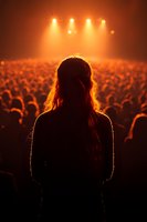 Silhouette of woman with long hair facing large crowd at concert venue with dramatic orange stage lighting and atmospheric smoke for entertainment events.